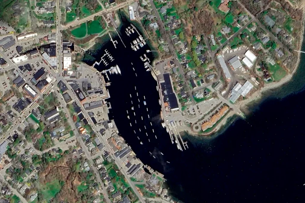 A high-resolution satellite image of a coastal town with a harbor filled with boats. The town features a mix of commercial and residential buildings, roads, and green spaces. The waterfront has multiple docks and marinas, with piers extending into the dark blue waters. The layout shows a dense downtown area on the left and a more suburban neighborhood with tree-lined streets on the right. The shoreline is well-developed, with some industrial or storage facilities near the water.