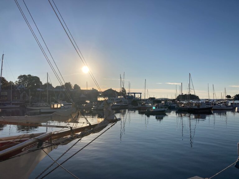 A peaceful harbor scene at sunrise or sunset, with numerous sailboats and motorboats docked in calm waters. The sun is low in the sky, casting a warm glow over the boats and reflecting on the water. Rigging lines from a nearby sailboat in the foreground create diagonal lines, adding depth to the composition. In the background, small buildings and trees line the shore, contributing to the serene coastal atmosphere.